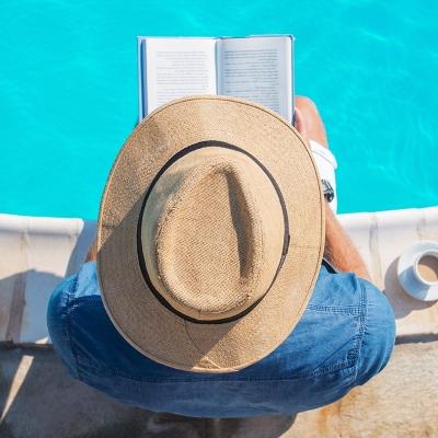 person wearing hat, reading book and sitting by pool with legs in the water